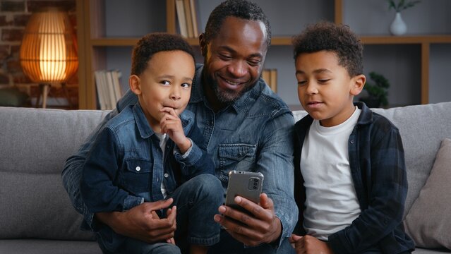Happy African American family looking in smartphone at home father with two little boys children kids enjoying mobile phone app social media internet smiling watching funny video on couch talking