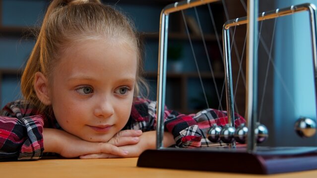 Curious little girl child kid daughter watching Newton cradle balls swinging on desk learning physics experiment at home in evening thoughtful observing motion science education curiosity in childhood