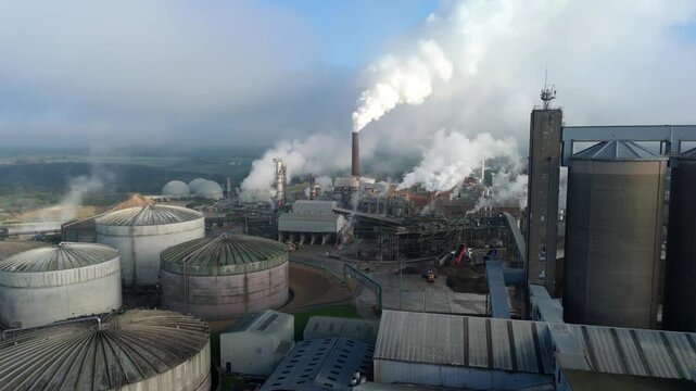 Aerial view of a factory complex with huge tanks and a tall chimney expelling smoke, contrasting with the surrounding landscape, Bury Saint Edmunds, England, United Kingdom.