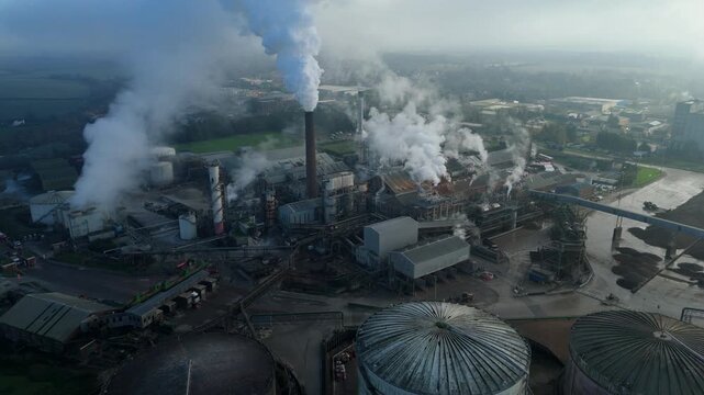Aerial view of a sprawling factory emitting plumes of white smoke into the hazy sky, contrasting against the green fields, Bury Saint Edmunds, England, United Kingdom.