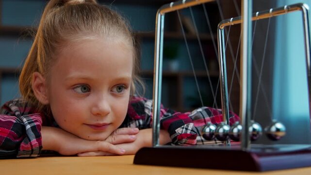 Curious little girl child kid daughter watching Newton cradle balls swinging on desk learning physics experiment at home in evening thoughtful observing motion science education curiosity in childhood