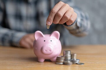 Man deposits a coin into a pink piggybank surrounded by stacks of coins on a wooden table