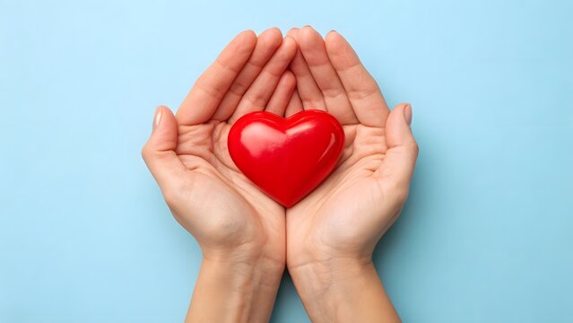 Human hands tenderly holding a bright red heart against a soft blue background, embodying concepts of love, health, care, charity, donation, and global wellbeing