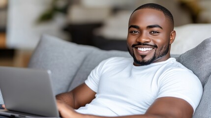 A smiling African American man with a beard is using a laptop while relaxing on a grey couch with pillows in a living room at his Home
