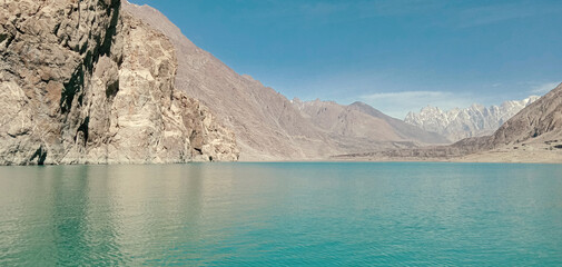 The image captures the calm, expansive turquoise water of a mountain lake meeting massive, rugged slopes on all sides. Distant snow-capped peaks anchor the horizon, showcasing the dramatic scale where