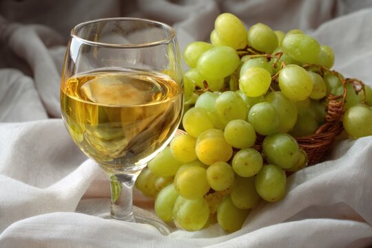 Elegant glass of white wine placed next to a cluster of fresh green grapes on a soft white fabric background