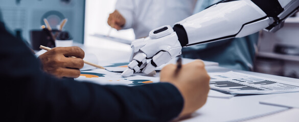 Human and robot collaborate during a meeting, analyzing data charts on a table, showcasing the integration of technology and artificial intelligence in the business environment.Noogenesis