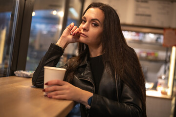 Woman with long dark hair, wearing a black leather jacket, sits at a wooden counter holding a coffee cup, exuding a confident and relaxed vibe in a cozy cafe environment
