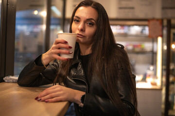 Woman with long dark hair, wearing a black leather jacket, sits at a wooden counter holding a coffee cup, exuding a confident and relaxed vibe in a cozy cafe environment