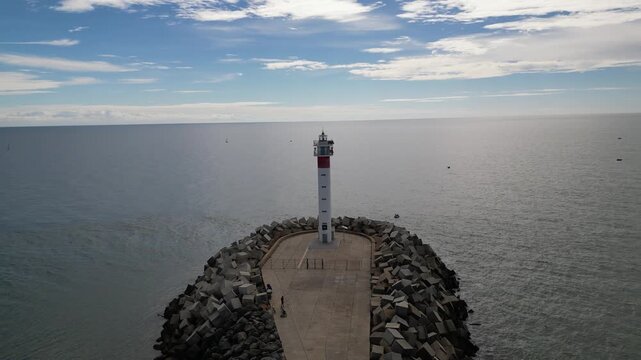 Aerial view of the striking red and white Faro de El Espigon lighthouse standing tall against the tranquil ocean waters under a partly cloudy sky, Huelva, Andalusia, Spain.