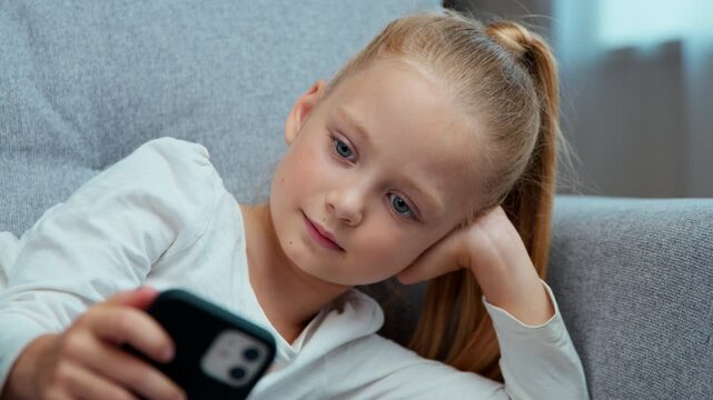 Relaxed Caucasian child little girl daughter kid teen with phone resting on couch in living room using mobile application modern technology online chat internet games at home with smartphone device