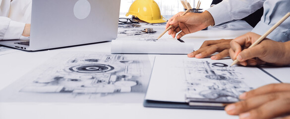 A modern office scene showing engineers collaborating over blueprints and designs while utilizing laptops. Hard hat and sketches highlight project planning and teamwork. SACTR