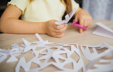 A child creating paper white snowflakes at a table at home. A child cuts patterns on paper with...