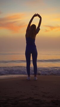 Young athletic woman in sportswear stretching her body on sandy beach, performing warm up exercises with her back to camera against stunning sunset over ocean