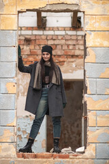 Obraz premium Young woman with long dark hair stands confidently in a broken wall opening, wearing a stylish black coat and beanie, showcasing urban decay and fashion in a gritty environment