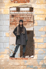 Young woman with long dark hair stands confidently in a broken wall opening, wearing a stylish black coat and beanie, showcasing urban decay and fashion in a gritty environment