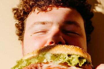 Young man with curly hair joyfully biting into an oversized sesame sandwich filled with fresh ingredients, capturing a moment of indulgence and happiness in a warm atmosphere