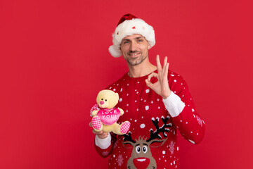 Santa with toy teddy bear. Portrait of middle aged man in sweater isolated over red background. Concept of holidays, happiness, emotions and Christmas celebration.