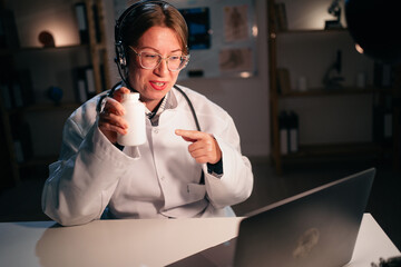 Female doctor or nurse in uniform wear headset use laptop computer talking on video call with patient recommend pills in hospital during night shift. Consulting, telemedicine and therapy