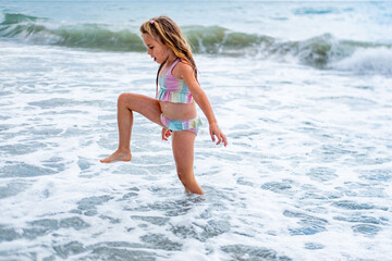 girl in colorful swimsuit jumping by sea shore, girl playing in the shallow water near seashore, waves warm summer day, family summer vacation kids splashing, enjoying carefree childhood play moments