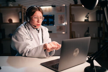 Female doctor in uniform wear headset gesture for kindness with help use laptop computer talking on video call with patient in hospital during night shift. Consulting, telemedicine and therapy