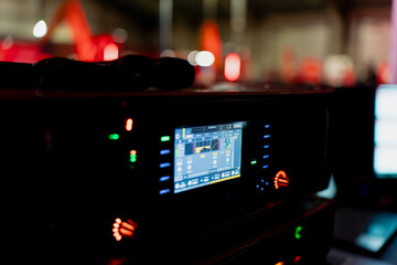 Electronic audio control unit with glowing interface, level indicators and active screen situated in dimly lit stage operations area during live event setup