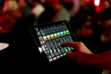 Obraz premium Tablet based audio mixer in a busy event space with soft red lighting, illustrating the technical side of live event production, sound management, and modern marketing driven audience experiences
