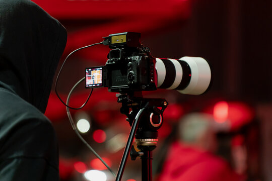 Tripod mounted camera with display screen and connected cables in a red illuminated venue, highlighting the technical setup behind event marketing coverage, filming, and live broadcast creation