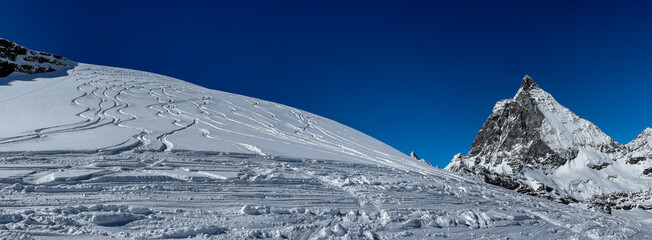 Panoramic winter landscape of the Matterhorn and surrounding alpine slopes in Switzerland, showcasing fresh ski tracks, bright blue sky, and prisitine mountain scenery.