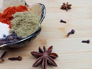 Close up of assorted spices including paprika, ground coriander, salt and black peppercorns in a decorative bowl, with star anise and cloves scattered on a wooden surface. Warm natural colors and deta