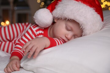Baby in Christmas pajamas and Santa hat sleeping on bed indoors