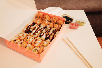 An open delivery box with assorted fried sushi rolls, soy sauce, ginger, wasabi, and chopsticks on a white table. A cozy interior setting highlights modern takeaway dining and Asian food culture