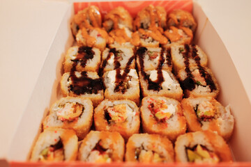 A close view of assorted fried sushi rolls arranged in a delivery box, topped with sauces and crispy textures. The image highlights variety, richness, and modern takeaway cuisine