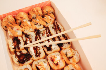 Chopsticks rest on a box of assorted fried sushi rolls topped with sauces and crispy textures. The...