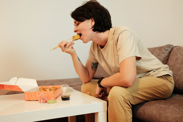 A woman sits on a sofa and eats fried sushi with chopsticks from a delivery box. The scene highlights casual dining, comfort, and modern home lifestyle with Asian food