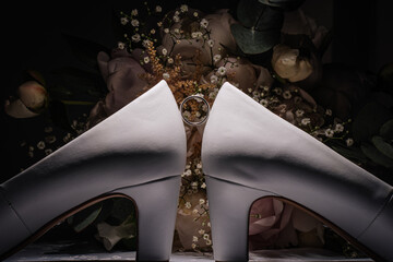 Elegant white high-heeled shoes positioned with a wedding ring between them, surrounded by a...
