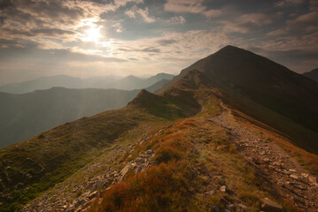 View of rugged mountain peaks under a sky streaked with clouds, where sunlight filters through, illuminating a path winding along the ridge, Pribylina, Zilina Region, Slovakia.