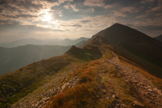 View of rugged mountain peaks under a sky streaked with clouds, where sunlight filters through, illuminating a path winding along the ridge, Pribylina, Zilina Region, Slovakia.