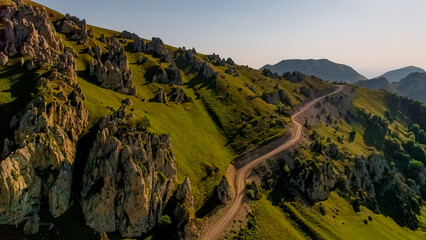 Aerial view of a winding road through green hills and rocky formations in the Caucasus Mountains. The landscape is illuminated by sunlight, showcasing natural beauty.