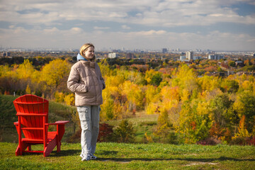 Fototapeta premium Young woman standing on a hill with hands in pockets, enjoying panoramic autumn views of Toronto skyline and colorful forest below