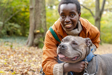 Black woman hugging large grey dog during autumn walk in forest park, medium close up
