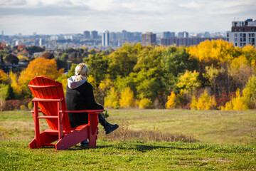 Fototapeta premium Middle-aged woman sitting on a red wooden chair atop a hill, overlooking colorful autumn trees and the distant Toronto skyline