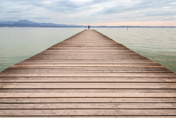 A long wooden pier extends into calm water under a soft, cloudy sky, with distant mountains on the horizon and a lone person standing at the end, creating a peaceful, minimalist scene.
