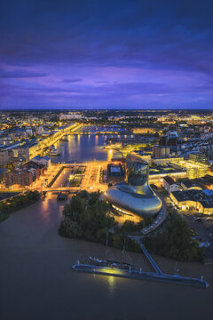 Aerial view of the Mus&eacute;e du Vin gleaming against the dimming twilight, its futuristic curves reflecting the city lights and the Garonne river, Bordeaux, Nouvelle-Aquitaine, France.