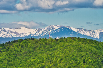 Aerial view of lush green hills contrasted against snow-capped mountains under a cloudy sky, Banska Bystrica, Banskobystricky kraj, Slovakia.