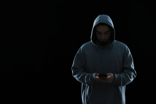 Criminal in hood with smartphone on black background, space for text