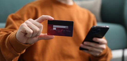 Young man making online payment with phone and credit card indoors, closeup