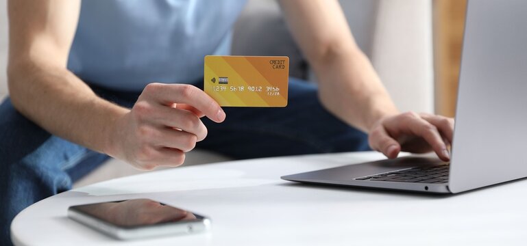 Young man making online payment with laptop and credit card at white table indoors, closeup