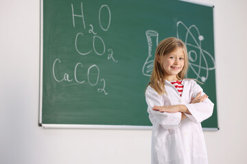 Smiling little girl in laboratory coat near green chalkboard with formulas indoors, space for text....