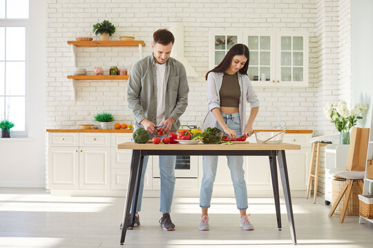 Young couple in love spends time together while preparing breakfast together in bright, cozy kitchen. Man and woman in casual clothes standing at wooden table and cutting vegetables for healthy salad.